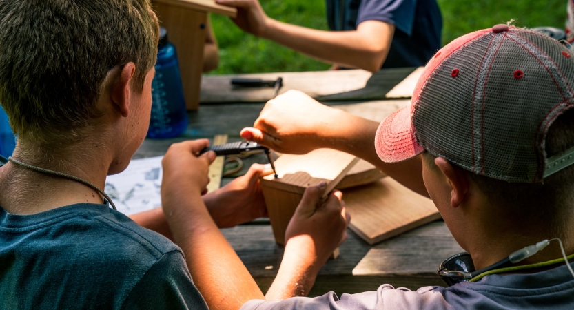 A young person holds two pieces of wood while another uses a tool to attach a screw
